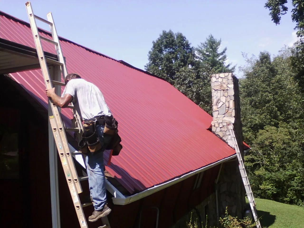 Roofer on a ladder beside a red metal roof and stone chimney — Soffit & Fascia Repair by Roofing Brothers in Edgerton